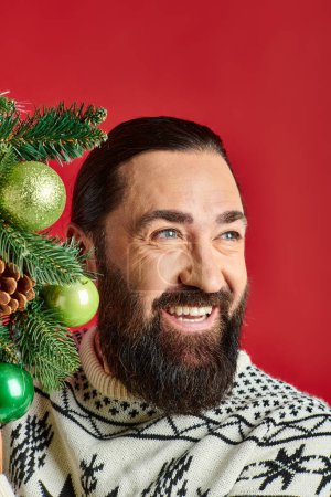 joyful bearded man in Christmas sweater holding decorated wreath with baubles on red backdrop