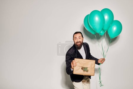 hombre barbudo feliz en traje sosteniendo globos azules y regalo de Navidad envuelto en fondo gris