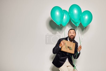 hombre barbudo alegre en ropa formal sosteniendo globos azules y regalo de Navidad sobre fondo gris