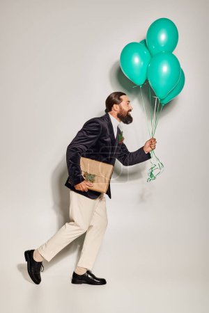 positive bearded man in formal wear holding blue balloons and walking with Christmas present on grey