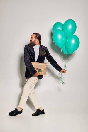happy bearded man in formal wear holding blue balloons and walking with Christmas present on grey