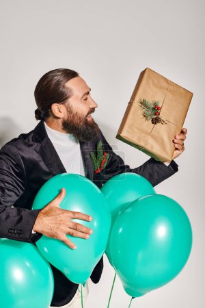 Foto de Sonriente hombre barbudo en ropa formal sosteniendo regalo de Navidad cerca de globos azules sobre fondo gris - Imagen libre de derechos
