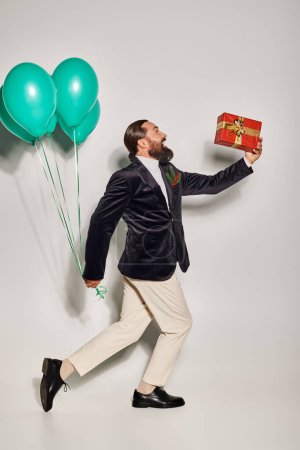 fröhlicher bärtiger Mann in festlicher Kleidung mit Weihnachtsgeschenk, der mit Luftballons vor grauem Hintergrund spaziert