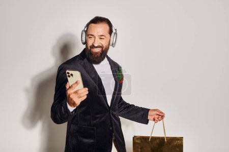 happy bearded man in headphones and suit holding smartphone and shopping bag on grey backdrop