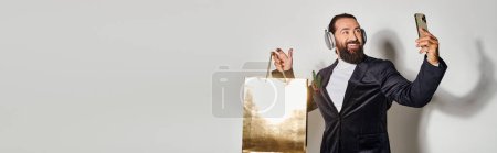 Photo for Happy bearded man in headphones taking selfie on smartphone and shopping bag on grey, banner - Royalty Free Image