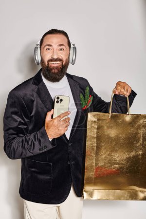 happy bearded man in headphones and suit holding smartphone and shopping bag on grey backdrop