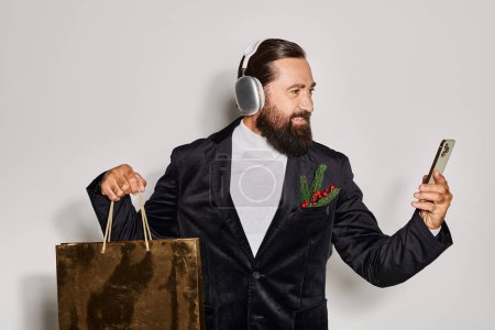 hombre barbudo feliz en auriculares inalámbricos que sostienen el teléfono inteligente y la bolsa de compras en el fondo gris
