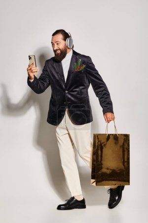 happy bearded man in wireless headphones holding smartphone and gift bag on grey, video call