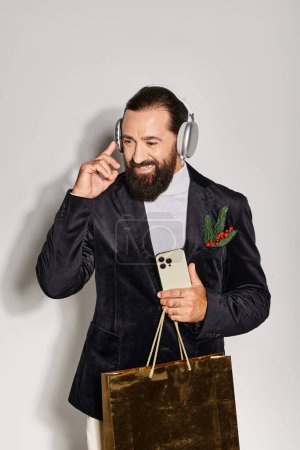 hombre feliz escuchando música en auriculares inalámbricos, sosteniendo teléfono inteligente y bolsa de regalo en el fondo gris