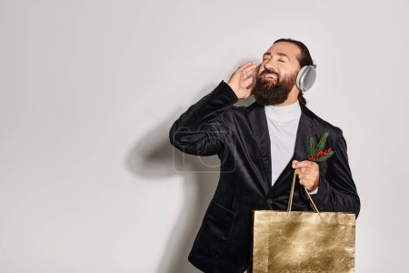 fröhlicher Mann, der Musik in drahtlosen Kopfhörern hört, Smartphone und Geschenktüte vor grauem Hintergrund hält