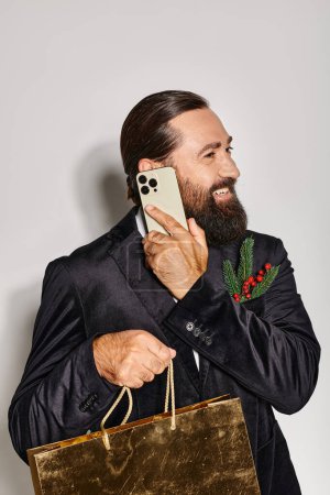 Photo for Happy bearded man in suit talking on smartphone and holding Christmas gift bag on grey backdrop - Royalty Free Image