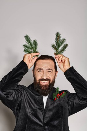 happy bearded man in suit holding two spruce branches near head and smiling on grey backdrop