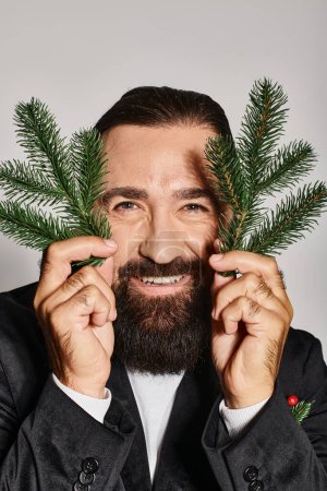 happy bearded man in suit holding two spruce branches near face and smiling on grey backdrop