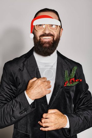 happy bearded man in festive glasses with santa hat posing in suit and smiling on grey backdrop