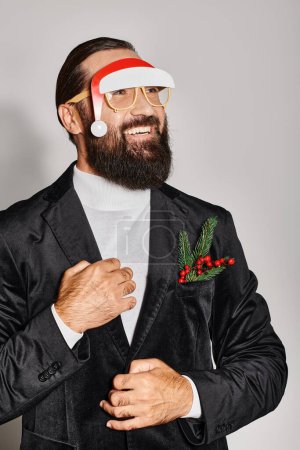 cheerful bearded man in festive glasses with santa hat posing in suit and smiling on grey backdrop