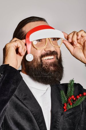 glücklicher bärtiger Mann in festlicher Brille mit Weihnachtsmütze posiert im Anzug und lächelt vor grauem Hintergrund