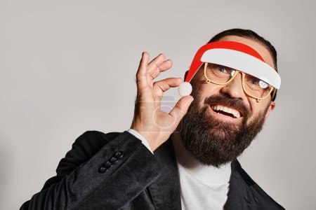 excited bearded man in festive glasses with santa hat posing in suit and smiling on grey backdrop