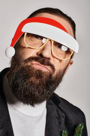 bearded man in festive glasses with santa hat posing in suit and pouting lips on grey backdrop