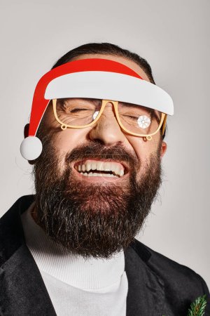Foto de Hombre barbudo alegre en gafas de fiesta con sombrero de santa posando en traje y sonriendo sobre fondo gris - Imagen libre de derechos
