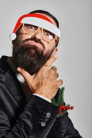 bearded man in festive glasses with santa hat posing in suit and pouting lips on grey backdrop