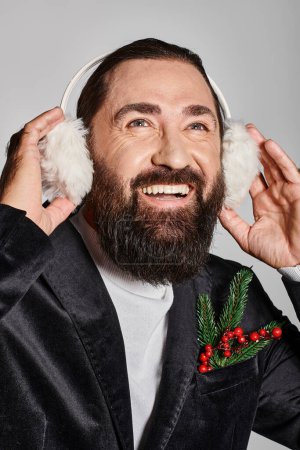 happy bearded man in suit with Christmas spruce branch and red berries wearing ear muffs on grey