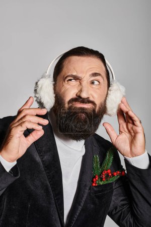 emotional bearded man in suit with Christmas spruce in pocket wearing ear muffs on grey backdrop