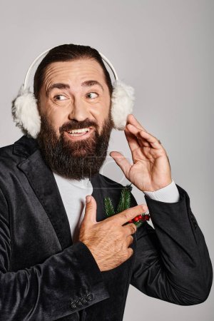 cheerful bearded man in suit with Christmas spruce in pocket wearing ear muffs on grey backdrop