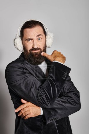 doubtful bearded man in suit with Christmas spruce branches and ear muffs moving eyebrows on grey