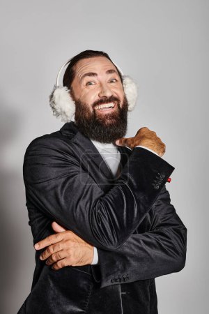 happy bearded man in suit with Christmas spruce branch and red berries posing in ear muffs on grey