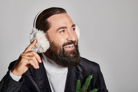 cheerful bearded man in suit with Christmas spruce branch and red berries wearing ear muffs on grey