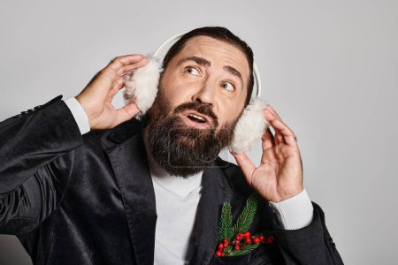 distracted bearded man in suit with Christmas spruce branches touching ear muffs on grey backdrop