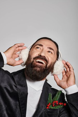 joyful bearded man in suit with Christmas spruce branches touching ear muffs on grey backdrop
