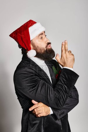 portrait of bearded handsome man in formal wear and santa hat posing on grey backdrop, Christmas