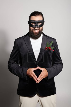 Masquerade ball, cheerful bearded man in carnival mask and elegant formal wear on grey backdrop