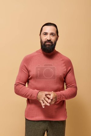 hombre barbudo guapo en jersey casual cuello alto mirando a la cámara en fondo beige, moda masculina