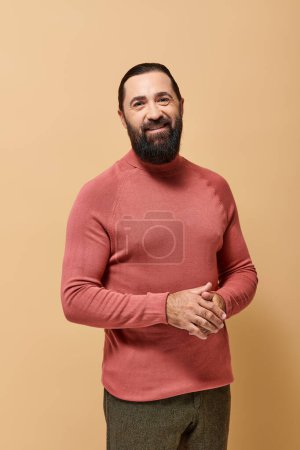 sonriente hombre barbudo en jersey casual cuello alto mirando a la cámara sobre fondo beige, moda masculina