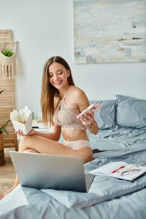 jolly woman in lingerie sitting on bed holding noodles and phone and smiling at her laptop camera