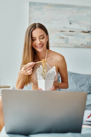 cheerful young woman eating noodles with chopsticks and looking at her laptop while working hard