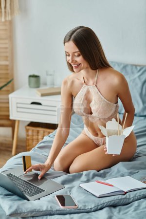 cheerful young woman in lingerie holding box of noodles and paying by credit card on internet