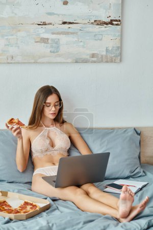 Foto de Mujer joven reflexiva en lencería con gafas sosteniendo rebanada de pizza mientras trabaja en su computadora portátil - Imagen libre de derechos