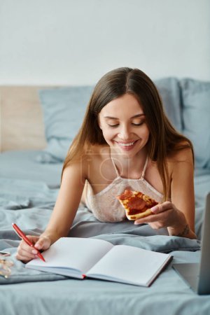 appealing cheerful woman in lingerie lying in bed enjoying pizza and taking notes while working hard