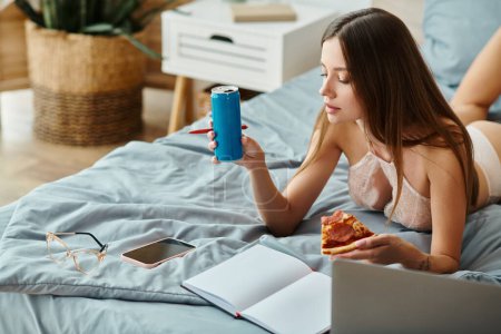 nachdenkliche junge Frau in sexy Dessous, die mit Pizza und Limo im Bett liegt, während sie aus der Ferne arbeitet
