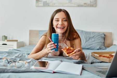 cheerful alluring woman with long hair lying on bed with pizza and soda and looking at camera