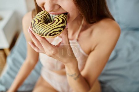 cropped view of young woman in sexy beige lingerie enjoying sweet donut while sitting on bed