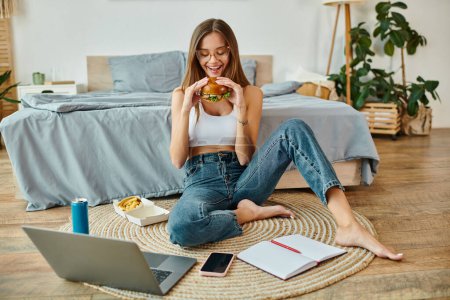 freudige schöne Frau in lässigem Outfit sitzt auf dem Boden und isst Burger, während sie von zu Hause aus arbeitet