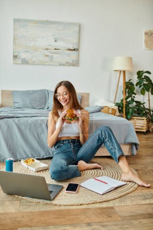 jolly young woman with glasses in cozy attire sitting on floor with burger and looking at her laptop