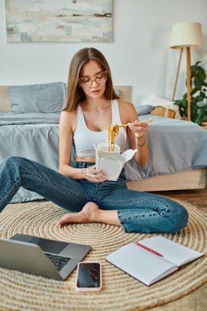Photo for Thoughtful beautiful woman in cozy homewear eating noodles while working remotely on laptop - Royalty Free Image