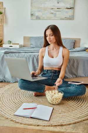 Photo for Beautiful young woman with long hair sitting with legs crossed eating popcorn while working remotely - Royalty Free Image