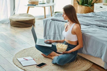 Photo for Attractive woman in comfy homewear sitting on floor and working at her laptop with popcorn in hand - Royalty Free Image