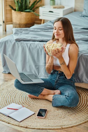 Photo for Cheerful young woman in homewear with long hair with laptop on her legs enjoying fresh popcorn - Royalty Free Image